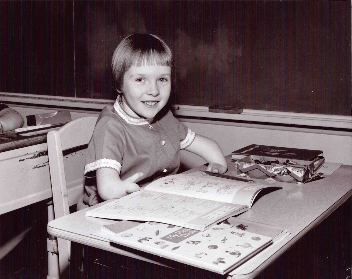 A black and white picture of a girl with shortened arms and missing fingers sitting at a school desk in the 1960s.